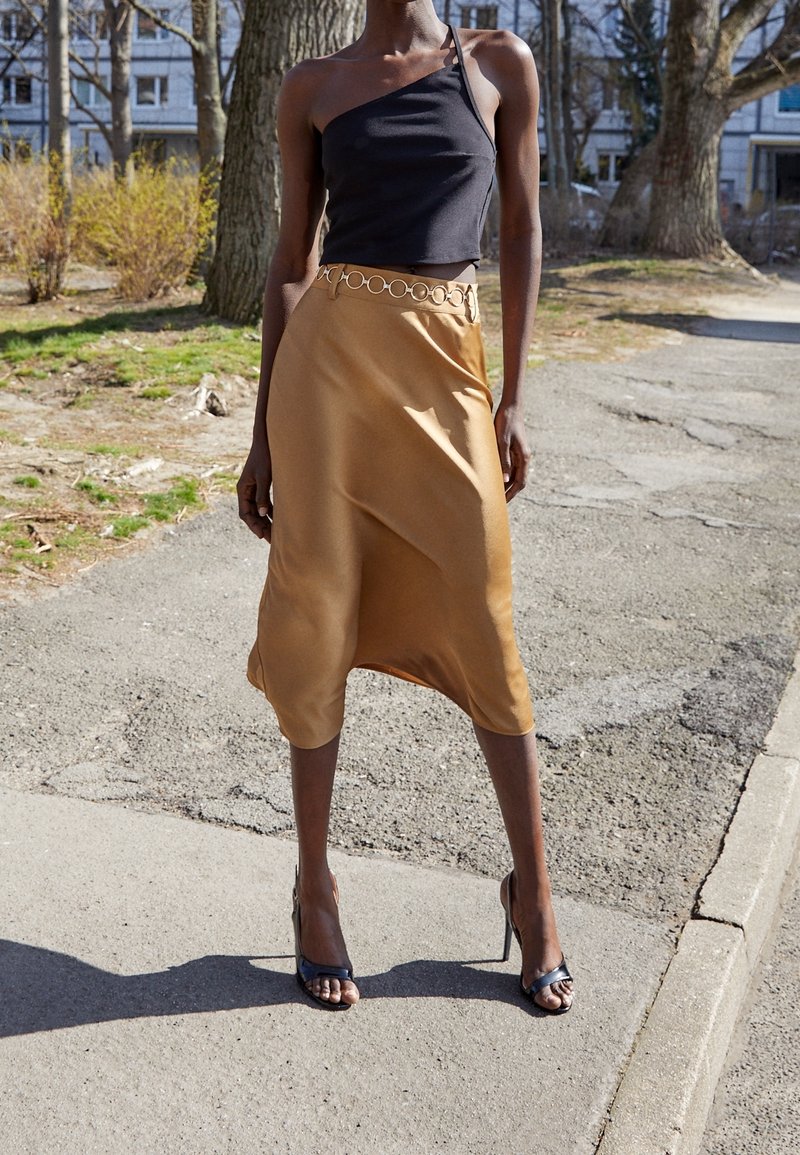 Femme debout en plein air, portant un haut noir à une épaule, une jupe midi couleur beige avec une ceinture circulaire, et des sandales à talons noirs sur le trottoir.