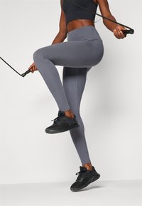 Grå leggings laget av elastisk stoff, høyt livdesign, kombinert med svarte treningssko, og holder en hoppetau.