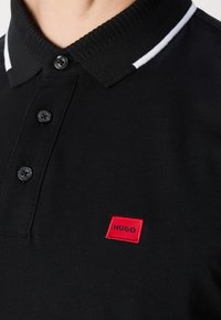 Svart polotröja med strukturerad tygkvalitet, med krage, tre svarta knappar och en röd rektangulär logotyp. Vit randig accent.