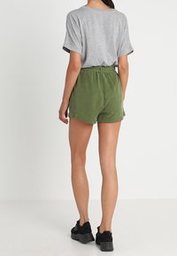 Šedé tričko s krátkým rukávem v kombinaci s vysokými zelenými šortkami. Šortky mají volný střih a hladkou texturu. Černé tenisky dokončují vzhled.