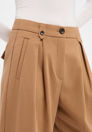 Pantalon beige ajusté avec plis, doté de deux boutons noirs et de poches latérales. Le tissu présente une texture lisse et un design structuré.