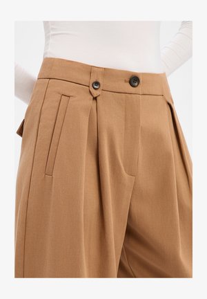Pantalon beige ajusté avec plis, doté de deux boutons noirs et de poches latérales. Le tissu présente une texture lisse et un design structuré.