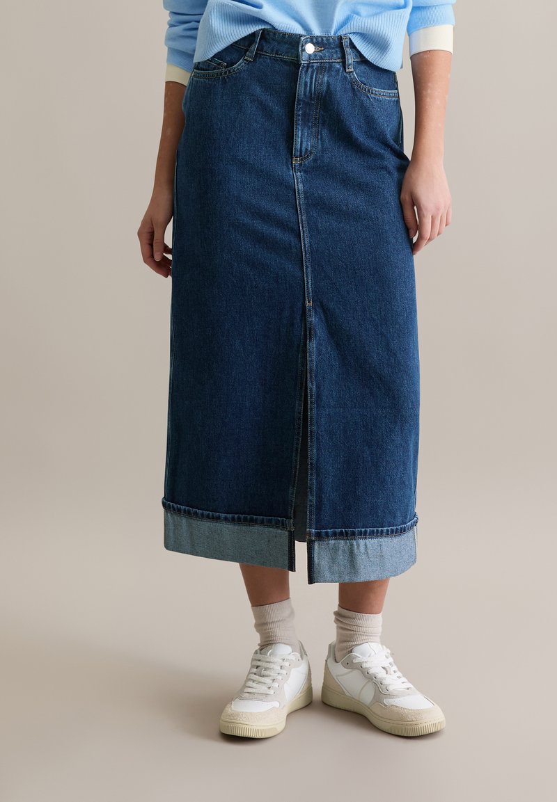 Długa spódnica dżinsowa w ciemnym niebieskim kolorze z przednim rozcięciem, z jaśniejszym, dżinsowym obszyciem, zestawiona z białymi trampkami i beżowymi skarpetkami.
