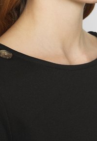 Černý top s kulatým výstřihem, hladkým povrchem a dekorativním knoflíkem na rameni. Materiál vypadá měkce a lehce.