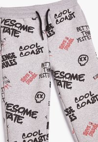 Graue Sweatpants mit schwarzem und rotem grafischem Text, der eine Vielzahl von Phrasen, ein Totenkopf-Motiv und einen Kordelzugbund zeigt.