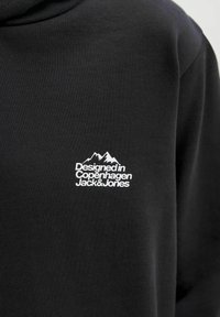 Čierna mikina so žltým textom loga s nápisom "Navrhnuté v Kodani Jack & Jones" pod grafickou ilustráciou hory na oblasti hrudníka.