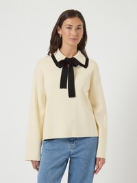 Mulher vestindo um suéter creme com colarinho e laço pretos, combinado com jeans azuis, de pé contra um fundo branco liso.