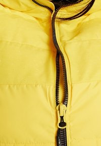 Gelbe Winterjacke mit glatter Textur, ausgestattet mit einem zentralen schwarzen Reißverschluss und gelbem Zugband. Sichtbare Abschnitte von genähten Steppnähten.