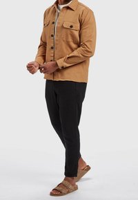 Kurtka zapinana na guziki w kolorze brązowym z dwoma kieszeniami na piersi, zestawiona z czarnymi zwężanymi spodniami i beżowymi sandałami. Gładka faktura, casualowy fason.