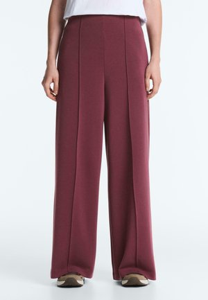 Osoba ubrana w wysokie, szerokie burgundowe spodnie z szerokimi nogawkami, w połączeniu z trampkami, stojąca na tle jednolitego jasnego tła.