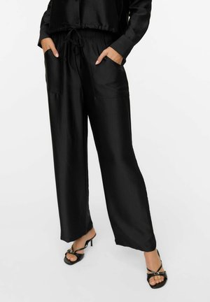 Pantaloni neri in satin con vestibilità comoda, vita elastica, due tasche laterali e texture liscia. Abbinati a sandali neri con tacco.