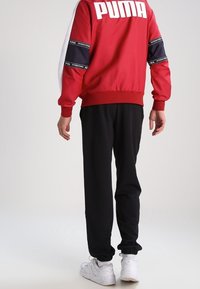Chaqueta deportiva roja con acentos en blanco y azul marino, con el logotipo "PUMA" en la parte posterior. Pantalones de chándal negros con puños elásticos y zapatillas blancas.