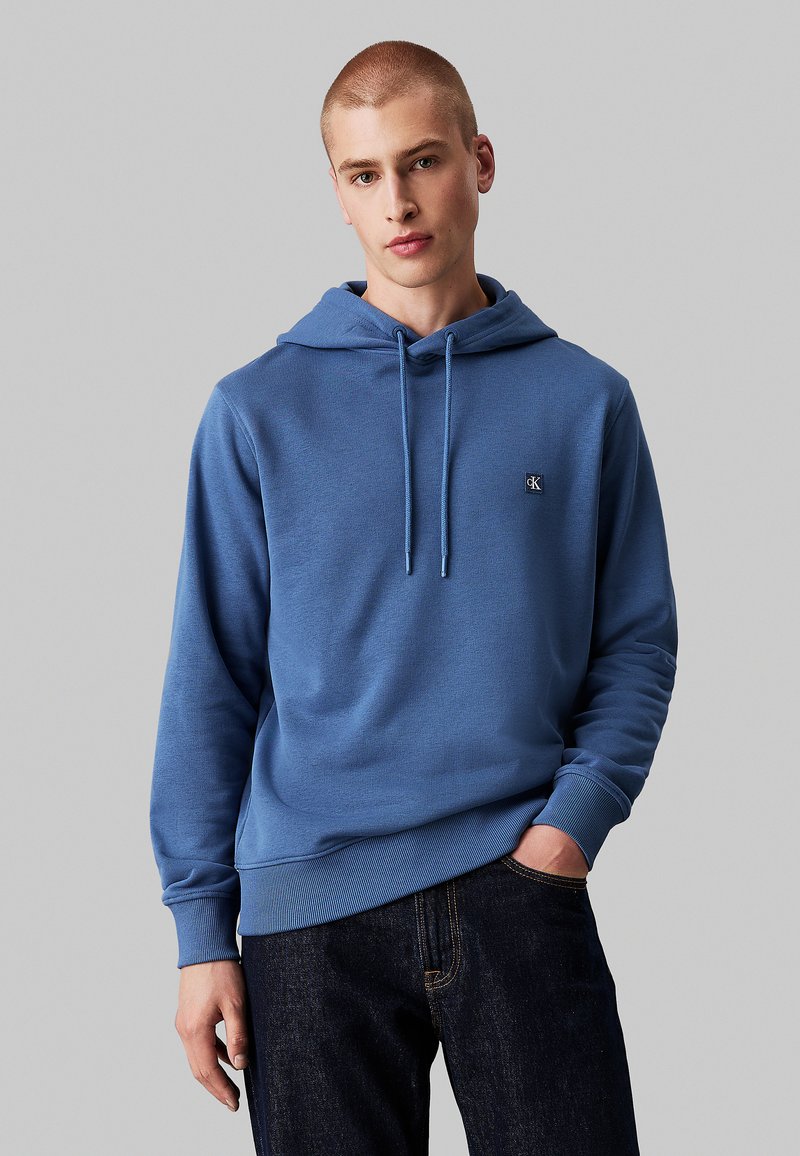 Felpa blu chiaro realizzata in tessuto testurizzato, con una toppa del logo, coulisse regolabile e polsini a costine. Abbinata a jeans scuri.