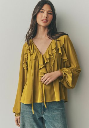 Blouse jaune moutarde avec un col et des manches à volants, dotée d'un décolleté en V et d'un détail à cordon, associée à un jean bleu.
