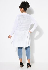 Lång vit cardigan med texturerad tyg, öppen rygg och plisserad design. Bärs över ljusblå tighta jeans och svarta högklackade skor.