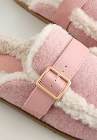 Lyserøde, fuzzy hjemmesko med en blød uldoverflade, fremhævet af en rosa læderrem med guldspænde og fluffy hvidt for.