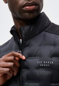 Κοντινό πλάνο ενός ατόμου που κλείνει το φερμουάρ ενός μαύρου καπιτονέ μπουφάν με το λογότυπο "TED BAKER SPORT" στο στήθος.