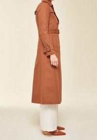 Hnědý trenčkot s ramenními epauletami, zlatými knoflíky, bočními kapsami a opaskem v pase, zkombinovaný s bílými širokými kalhotami a hnědými podpatky.