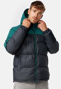 Padded jacket z górną częścią w kolorze szmaragdowym i dolną częścią w kolorze czarnym. Posiada kaptur, zamek błyskawiczny z przodu oraz boczne kieszenie z elastycznymi mankietami.