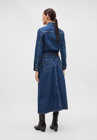 Jeanskjole med innsnevret midje, lange ermer og midilengde skjørt. Mørk blå farge, minimalistisk design og tradisjonell denimtekstur.