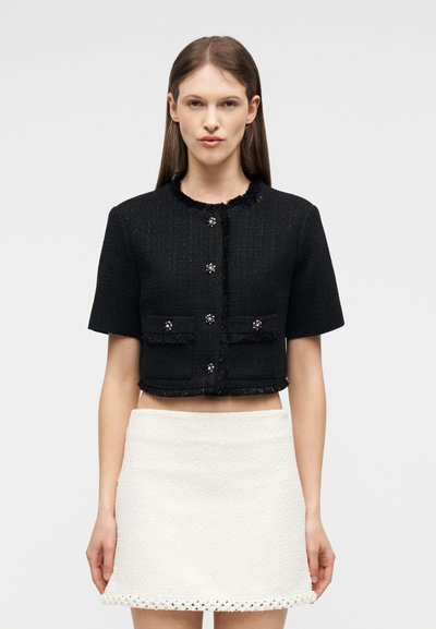 Γυναίκα που φοράει μαύρο κοντό μπουφάν με κοντά μανίκια και διακοσμητικά κουμπιά, συνοδευόμενο από λευκή υφασμάτινη μίνι φούστα με διακοσμητικό τελείωμα.