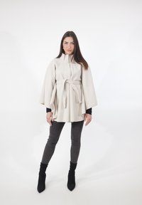 Beige cape-achtige buitenkleding met een hoge kraag, een ceintuur in de taille en vloeiende mouwen. Gecombineerd met donkergrijze skinny jeans en zwarte enkellaarsjes.
