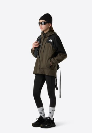 Veste imperméable verte et noire avec une fermeture éclair à l'avant, accompagnée de leggings noirs, d'un bonnet noir et de baskets sombres. Accents du logo blancs visibles.