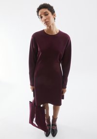 Bordeauxfarbenes Langarmkleid mit lockerer Passform, glatter Textur und minimalistischem Design. Begleitend dazu eine passende Tasche und schwarze Absatzschuhe.