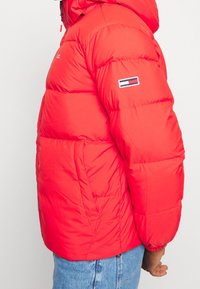 Rode pufferjack met capuchon, voorzien van gewatteerde secties, een ritssluiting aan de voorkant en een logopatch op de linkermouw. Geweven, geïsoleerde stof.