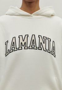 Biela mikina s kapucňou s nápisom "LAMANIA" v čiernych, tučných písmenách. Mäkká tkanina s klokaním vreckom a sťahovacou kapucňou. Minimalistický dizajn.