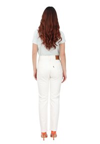 Pantalones de mezclilla blancos con cintura alta y pierna recta, que cuentan con bolsillos traseros y un parche de piel con marca. Modelo lleva tacones naranjas.