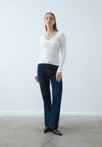 Maglietta a maniche lunghe, bianca a coste con scollo a V e dettagli con tre bottoni, abbinata a jeans blu scuro a gamba dritta e stivali neri alla caviglia.