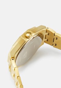 Gouden polshorloge met een geometrische kast, een gestructureerde kroon en een schakelarmband. Metallic afwerking met een combinatie van glanzende en matte oppervlakken.