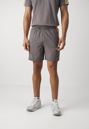 Mann trägt graue Adidas-Shorts mit gelben Streifen, weiße Adidas-Socken und weiße Sportschuhe, steht vor einem einfarbigen Hintergrund.