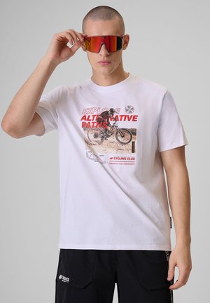 Osoba nosząca czerwone sportowe okulary przeciwsłoneczne z powłoką odblaskową, białą koszulkę z grafiką rowerową i czarne spodenki, stojąca na szarym tle.