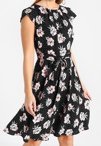 Czarna sukienka w kwiaty z krótkimi rękawami, ozdobiona różowymi kwiatami i kropkami, z talią podkreśloną szarfą oraz rozkloszowanym dołem.