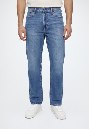 Homme portant un jean droit bleu moyen avec fermeture par bouton et fermeture éclair, associé à des baskets blanches et une chemise beige.
