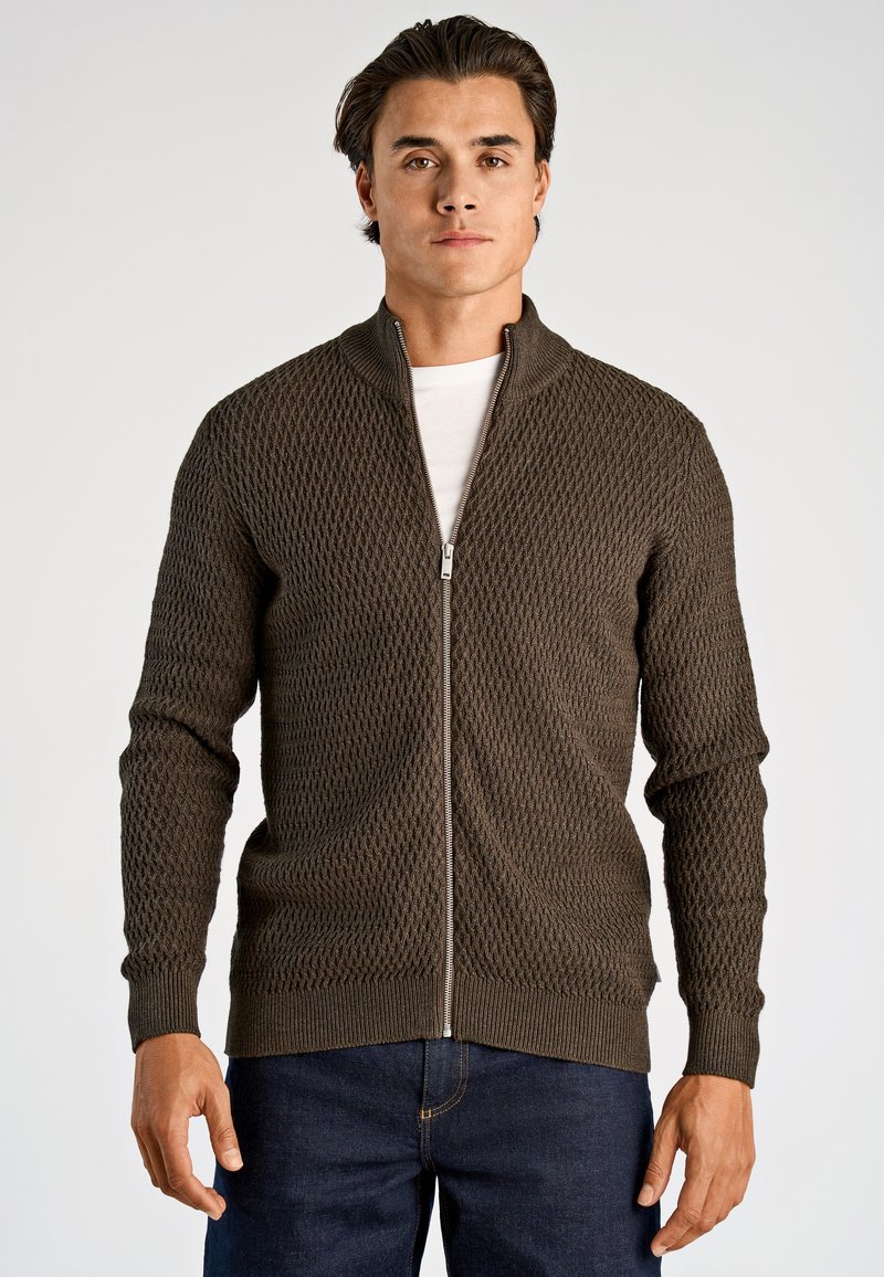 Homme portant un cardigan zippé texturé marron foncé sur une chemise blanche et un jean bleu foncé, debout devant un fond clair uni.