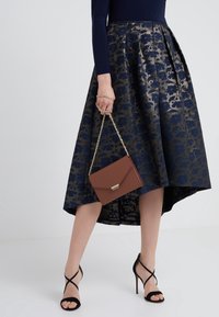 Tmavě modrá květinová sukně se zlatými akcenty, plisovaný design, lem s vysokým a nízkým střihem; hnědá kabelka se zlatým řetízkem, černé sandály na vysokém podpatku s pásky.