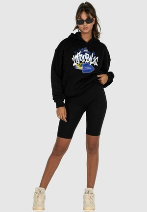 Schwarzer Hoodie mit grafischem Textdesign in Weiß- und Blautönen, kombiniert mit schwarzen Radshorts und beigen High-Top-Sneakern.