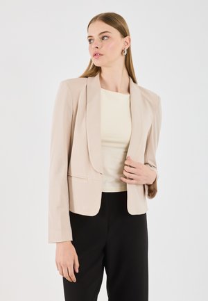 Joven mujer con blazer beige, top crema y pantalones negros, posando con una mano tocando el blazer, mirando ligeramente hacia su izquierda.