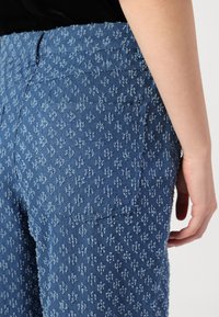 Blå denimbukser med et teksturert mønster og små blomstermotiver, detalj med baklomme og en figursydd design.