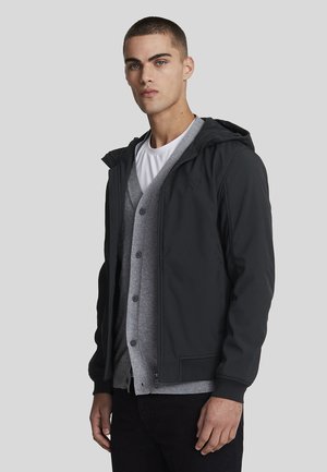 Sort letvægtsjakke med hætte, lynlåslukning og ribbede manchetter, lagdelt over en grå cardigan med knapper og en hvid t-shirt under.