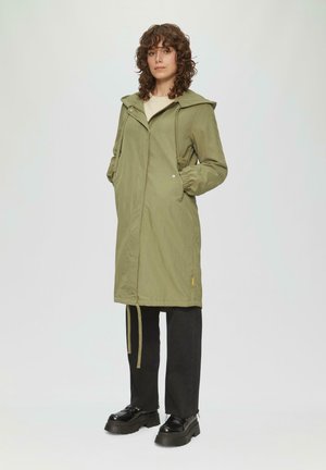 Olivgrüner, langer, kapuzenförmiger Regenmantel mit elastischen Bündchen, Reißverschluss und Kordelzug in der Taille. Getragen über schwarzen Hosen und klobigen schwarzen Schuhen.
