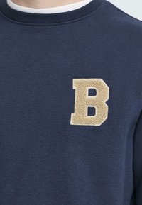 Navyblaues Sweatshirt mit Rundhalsausschnitt, das auf der linken Brust ein strukturiertes, beiges "B"-Applikation aufweist. Weicher Stoff mit glatter Oberfläche.