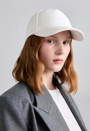 Gorra blanca de cuero con un diseño estructurado, que cuenta con una visera curva y detalles de costura, llevada con un blazer gris sobre una camisa blanca.