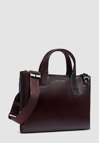 Bolso de cuero marrón con dos asas y correa desmontable. Presenta costuras azules y cierre de cremallera. Logotipo mostrado en la parte frontal.