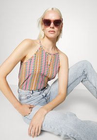 Top halter colorido com padrão zigzag em rosa, amarelo e azul, combinado com jeans de cintura alta azul claro. Óculos de sol rosa transparentes completam o visual.