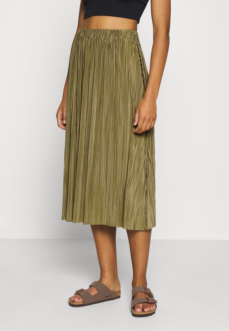 Femme portant une jupe midi plissée vert olive, un crop top noir et des sandales Birkenstock marron, debout devant un fond uni.