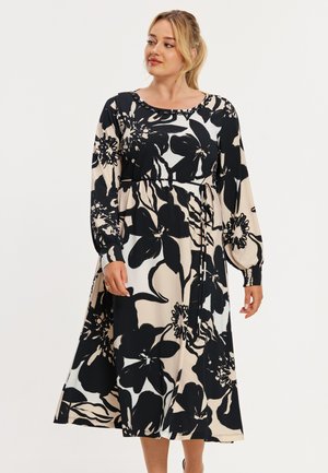 Kvinde iført en beige midi-kjole med store sorte blomstertryk, lange puffede ærmer og bæltet i taljen, stående foran en ensfarvet baggrund.